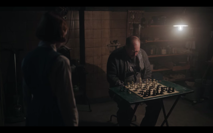 A man sits in front of a small table, which holds a chess board. He is looking down at the board. To the left, Beth is visible from behind as she watches him.