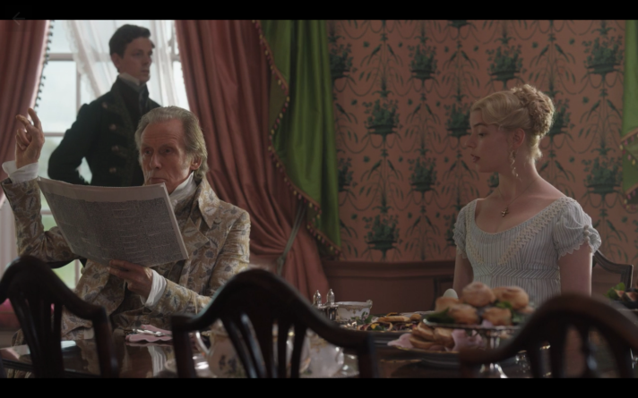 Emma's father, wearing a patterned jacket and white shirt with a high collar, sits next to Emma at the dining room table. He is reading a newspaper and has one hand raised as if he is about to silence Emma. A footman stands behind him in front of a window that his hung with thick pink and green curtains. The wall is papered with pink wallpaper that has a green design on it.