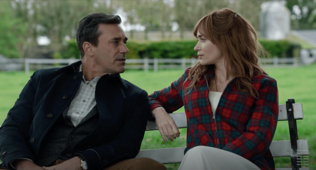 Jon Hamm, dressed in a black jacket, vest, and white checked button down shirt, sits on a bench next to Rosemary who has reddish long hair with bangs and is wearing a red plaid zip up barn coat over a flowing white dress.