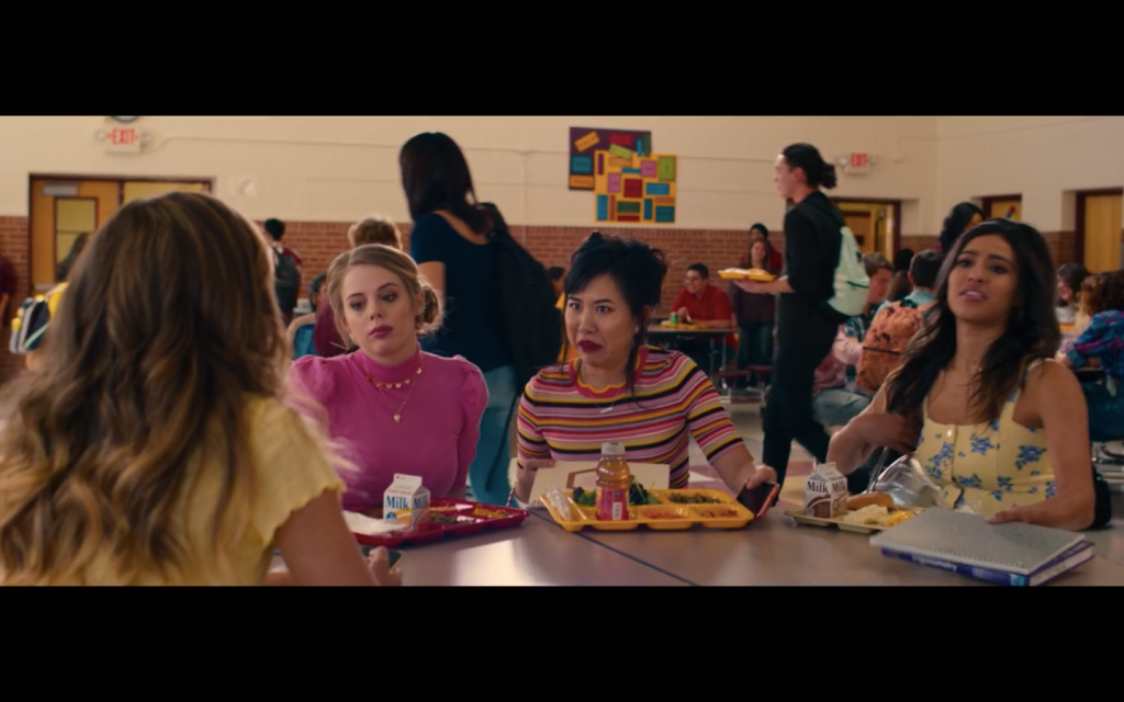 A blonde haired, white teenage girl in a hot pink shirt, sitting next to a black-haired asian teenage girl in a striped shirt, sitting next to a dark-haired black girl wearing a floral tank top. They are all seated at a table in a high school cafeteria.