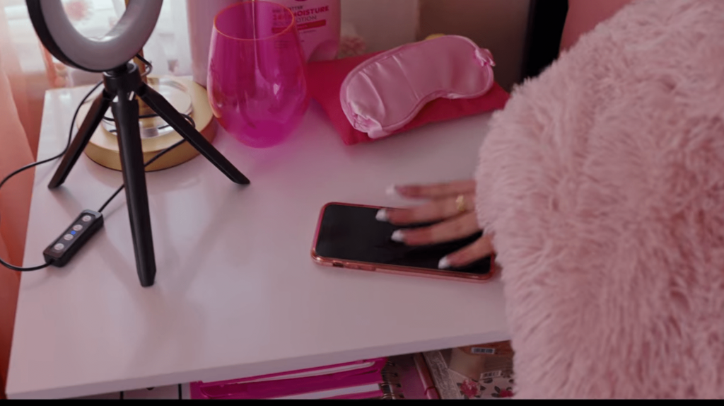 Padgett's hand on top of her phone on her nightstand. there is a ring light, a pink eye mask, a pink cup, a pink bottle of lotion, and a pink water bottle, her pink fleece blanket is also visible.