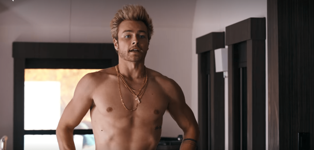 Jordan, with his blonde hair spike up high and wearing several gold chains, standing shirtless with his hands on his hips.