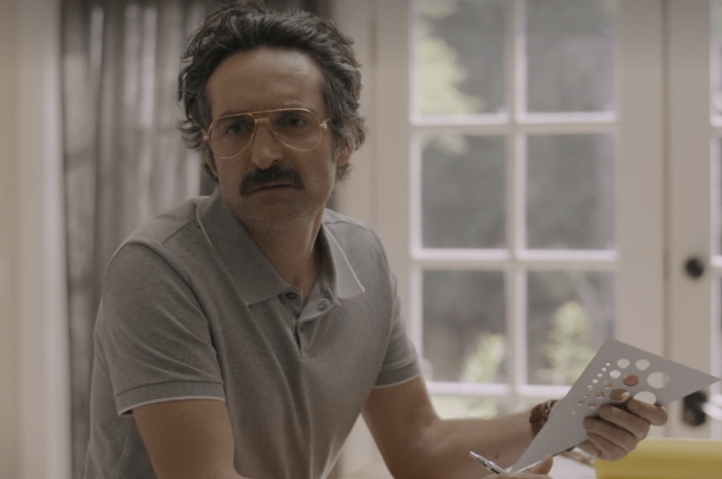 Martin, a white man with dark hair that reaches the nape of his neck. He is wearing gold aviator glasses and has a large mustache, narrow shoulders, and is wearing a grey polo shirt as he sits at his drafting table.
