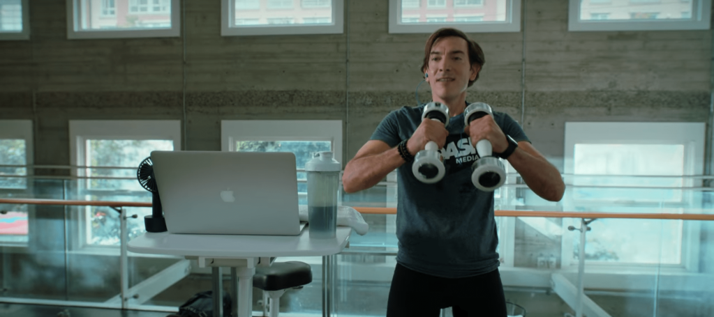 Lee standing next to his bike desk holding shake weights. He's wearing a t-shirt tucked into his bike shorts. He has short brown hair and is a white man.
