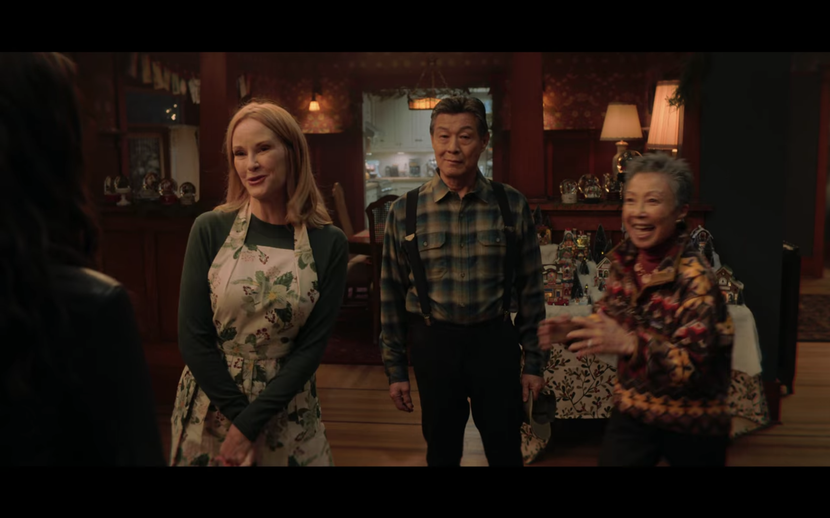 Josh's mom (a blonde white woman wearing an apron), Dad (an Asian man in a plaid shirt and suspenders), and grandmother (an older Asian woman with short grey hair and a colorful top) greeting Natalie.
