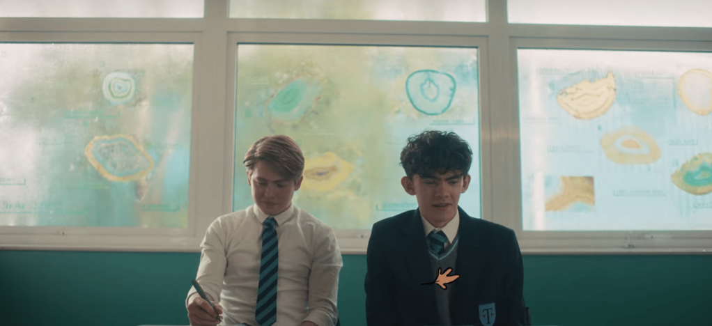 Nick and Charlie sitting next to each other in forms class. The half wall behind them is teal and then there are large square windows with yellow and blue shapes on them.