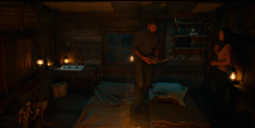 Max and Lola in a cabin standing over their bed rolls. Max is holding a green snake in his hands at waist level that has its head outstretched toward Lola.