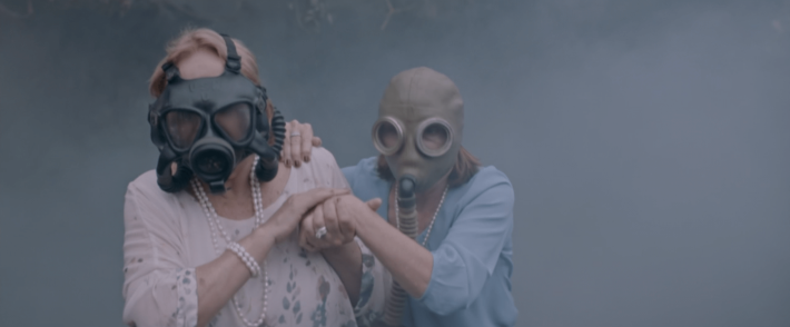 Alicia and Carmen wearing old-style gas masks as they move through a thick cloud a of tear gas. Both are still dressed in jewels and fine clothes for luncheon. Carmen has her hand on Alicias shoulder and her other hand is clasped in Alicia's as Alicia uses her free hand to pat Carmen's as they walk.s