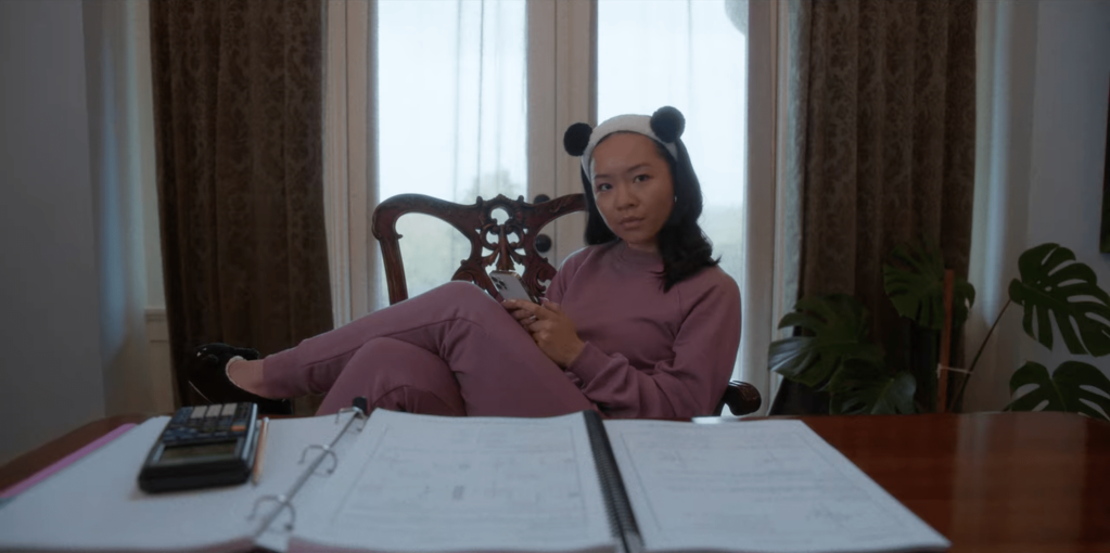 Becca in a pink sweatsuit and panda ears headband sitting at a glossy table with math homework in front of her. She hold her phone and is glaring at someone.