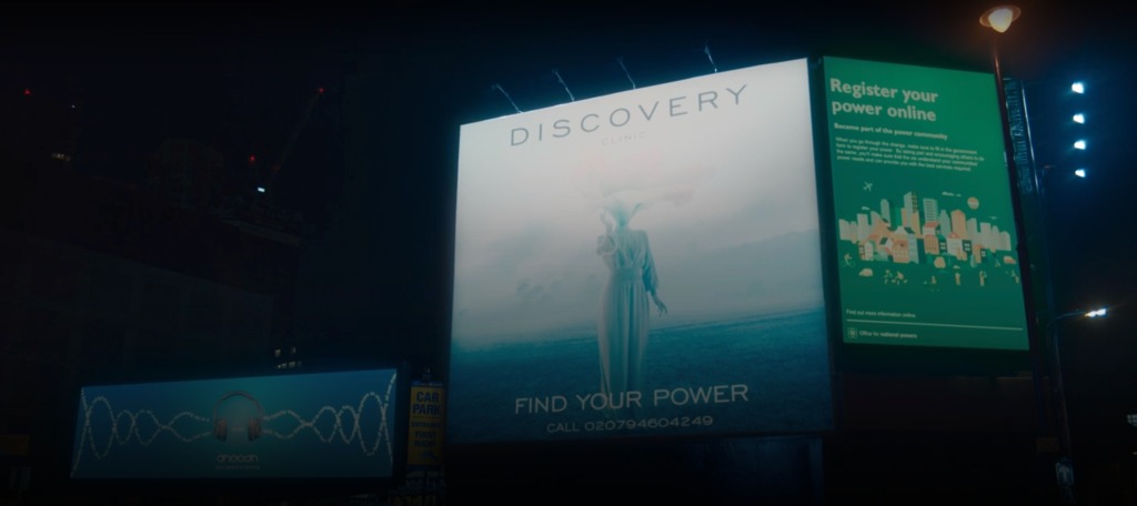 Several billboards. The central one is muted colors of off-white and blue. There is feminine person standing in the center with one hand raise. Their head appears to be an upside down lampshade? Text: Discover. Find your power. 
