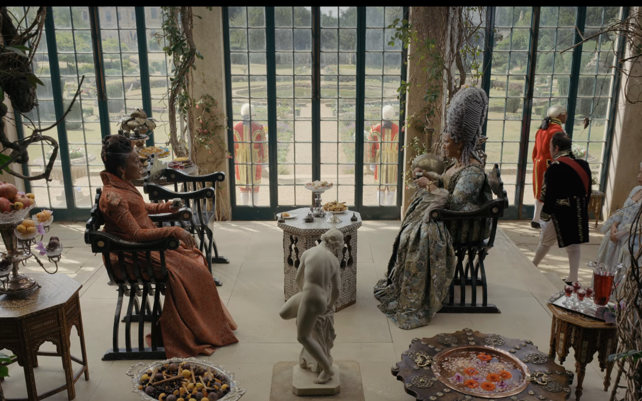 Lady Danbury and Queen Charlotte seated in wooden chairs in front of large glass doors as they have tea.